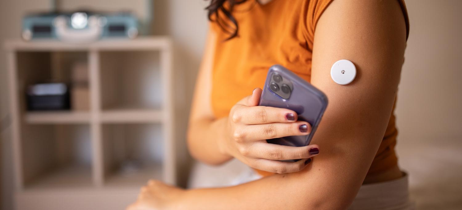 Woman holding a smartphone near a continuous glucose monitor sensor on her upper arm, representing remote health monitoring with wearable devices.