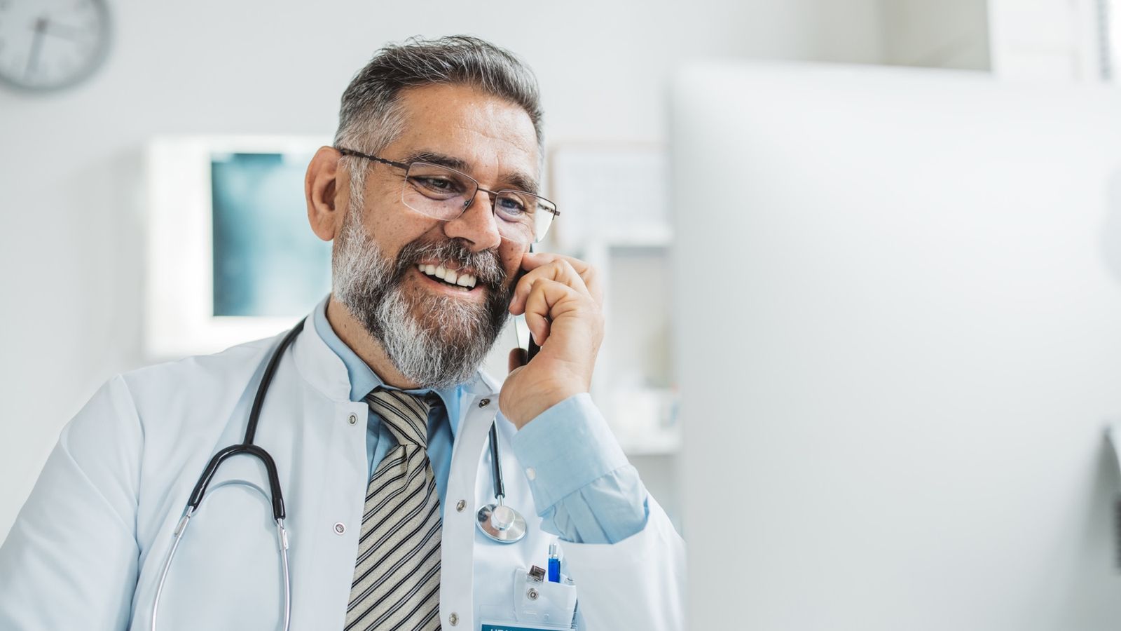 Doctor talking on a mobile phone in his office, smiling with a stethoscope and health center ID badge
