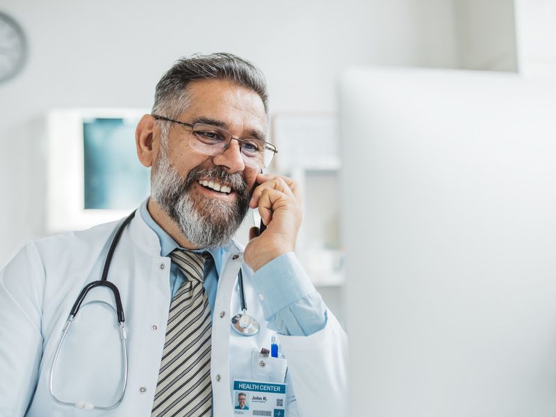Doctor talking on a mobile phone in his office, smiling with a stethoscope and health center ID badge