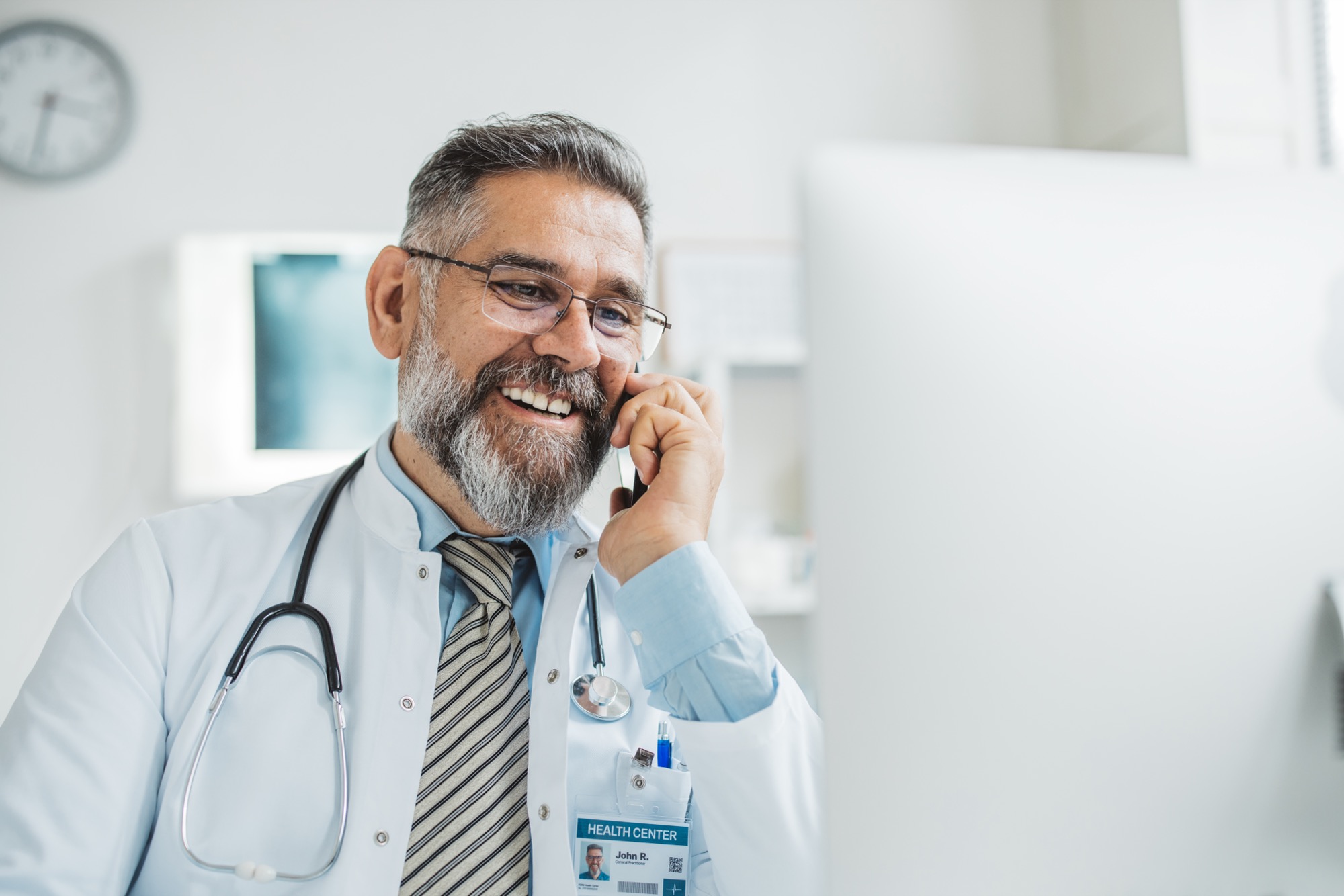 Doctor talking on a mobile phone in his office, smiling with a stethoscope and health center ID badge