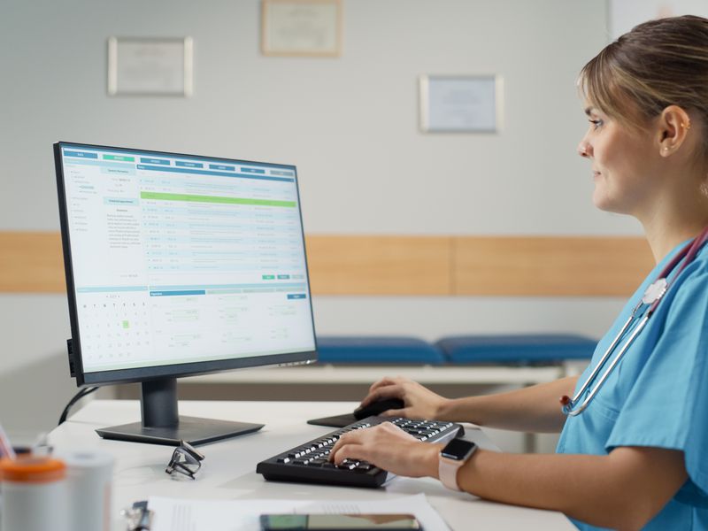 Nurse in blue scrubs reviewing electronic health records on a desktop computer monitor in a clinical setting.
