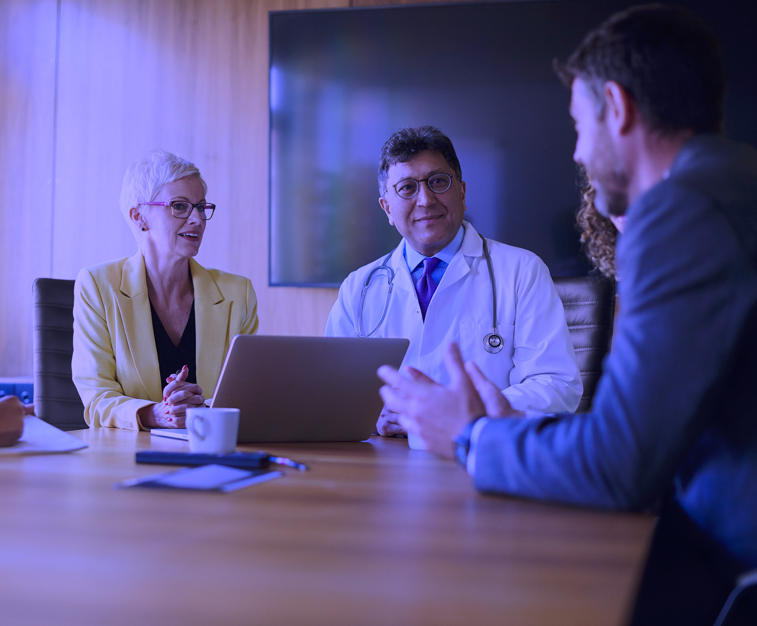 Healthcare administrators in a conference room meeting, including a doctor in a white coat and colleagues discussing around a laptop, with a purple color overlay.