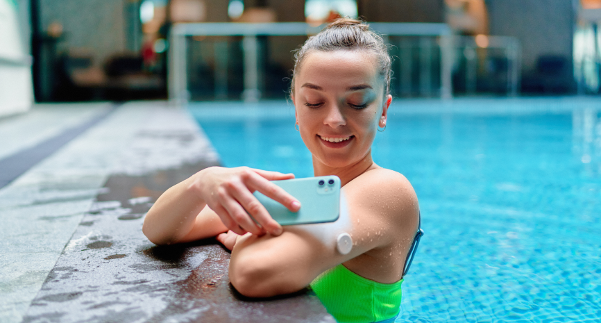 Woman smiling while using her smartphone at the edge of a swimming pool, wearing a fitness tracker on her wrist.