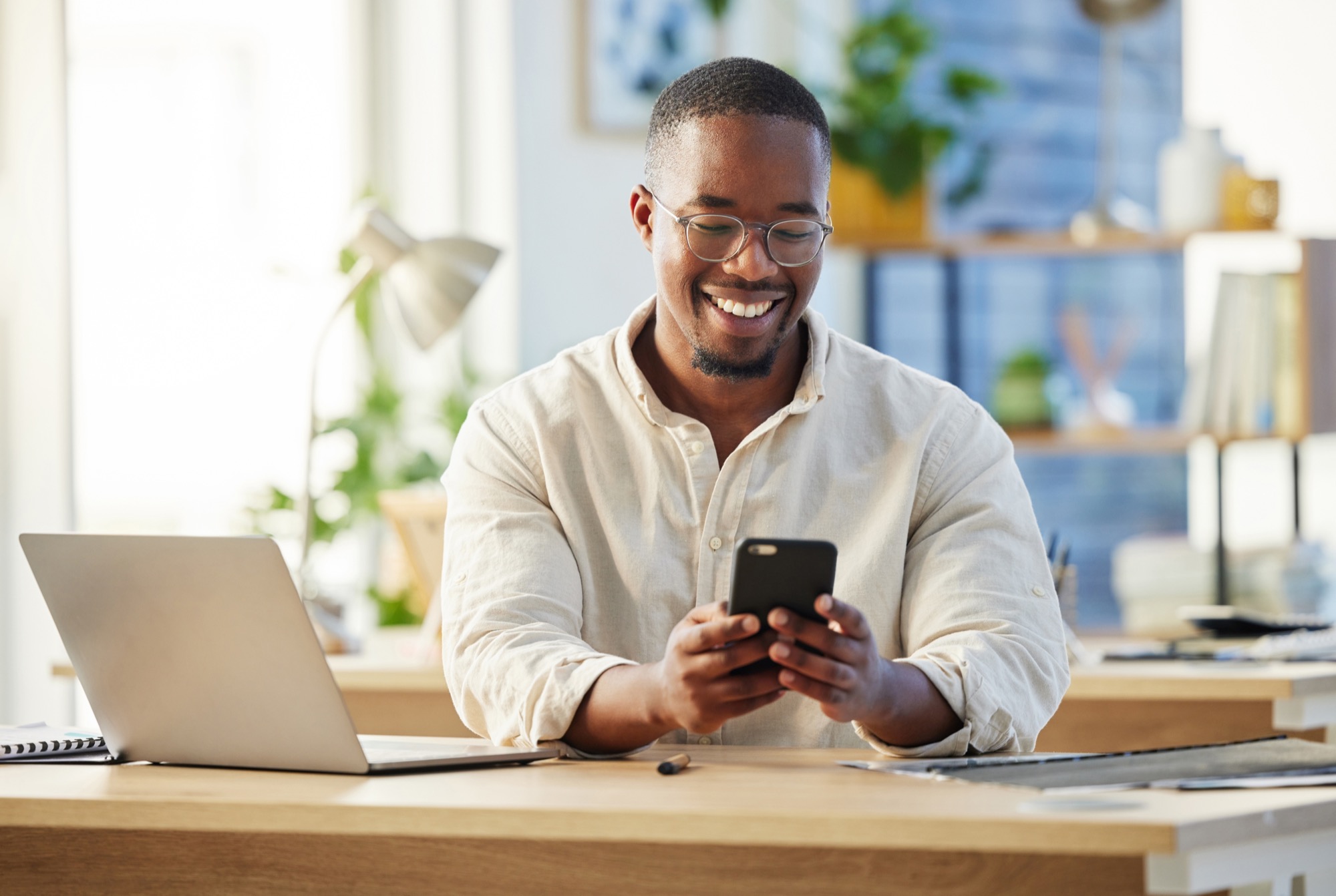 Man smiling while checking his smartphone at a desk with a laptop