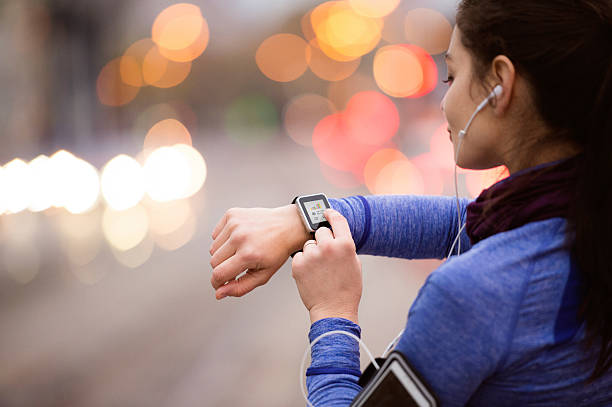 Female runner checking her smartwatch fitness tracker while wearing earbuds, with city lights blurred in the background.