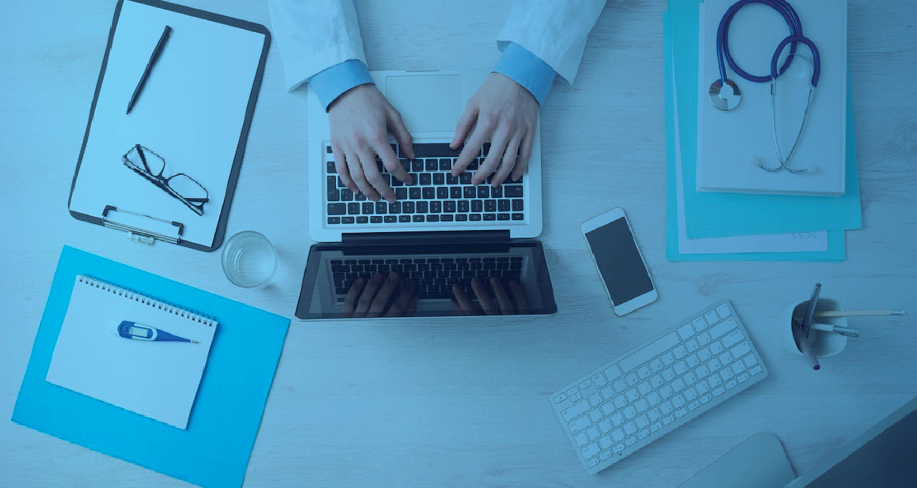 Overhead view of a clinician typing on a laptop at a desk with a stethoscope, smartphone, notepad, and medical thermometer.