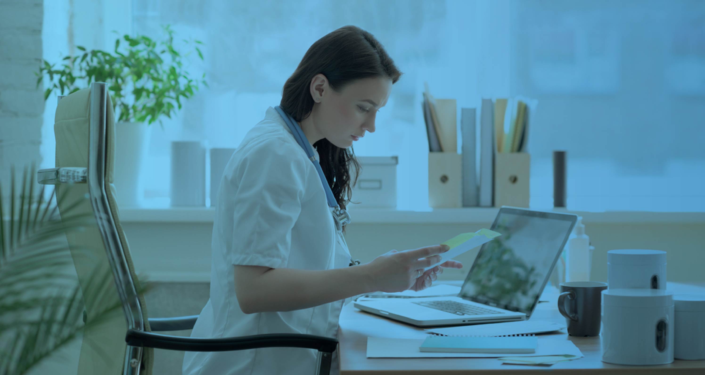 Healthcare professional in a white coat reviewing a document at her desk with a laptop, with a blue color overlay.