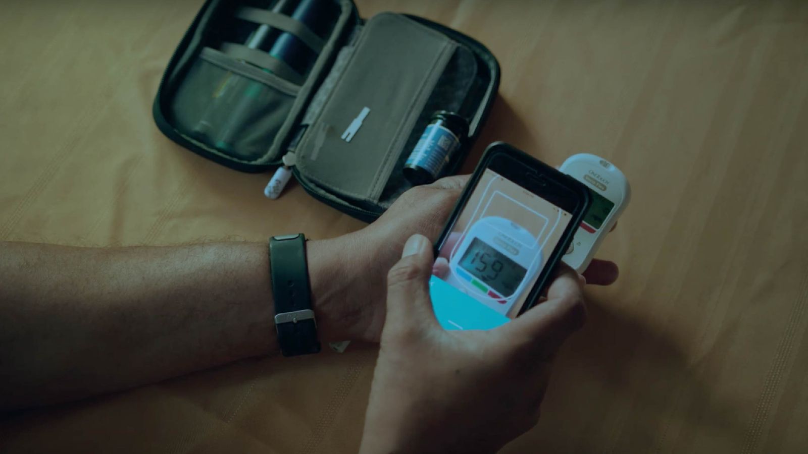 Person holding a smartphone displaying a blood glucose reading of 159 next to a glucose meter and diabetes testing kit, wearing a fitness tracker.