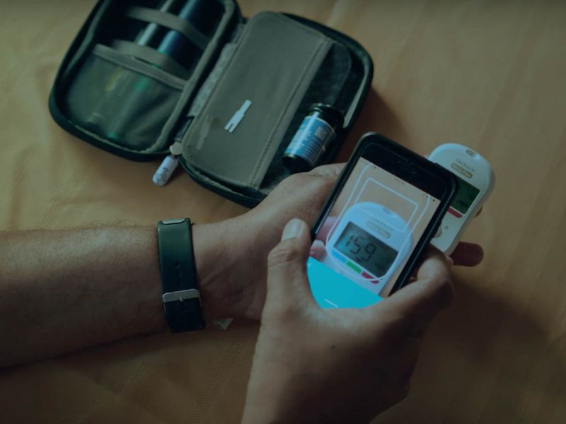 Person holding a smartphone displaying a blood glucose reading of 159 next to a glucose meter and diabetes testing kit, wearing a fitness tracker.