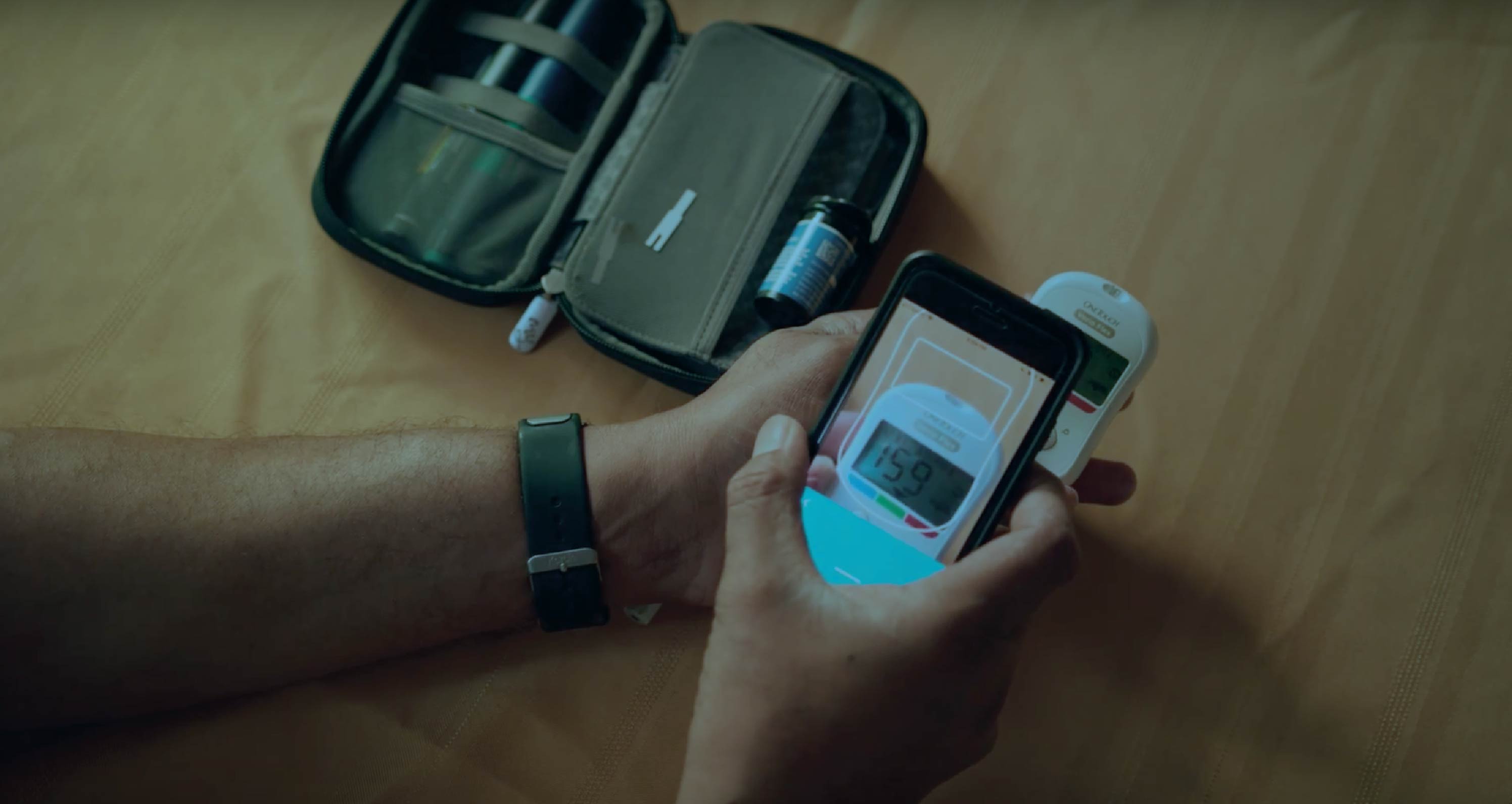 Person holding a smartphone displaying a blood glucose reading of 159 next to a glucose meter and diabetes testing kit, wearing a fitness tracker.