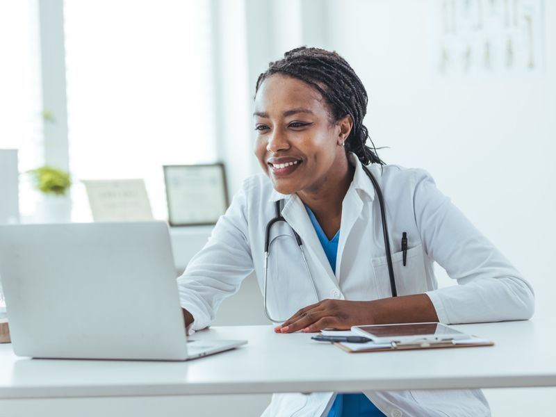 Female doctor with a stethoscope smiling while working on a laptop at her desk