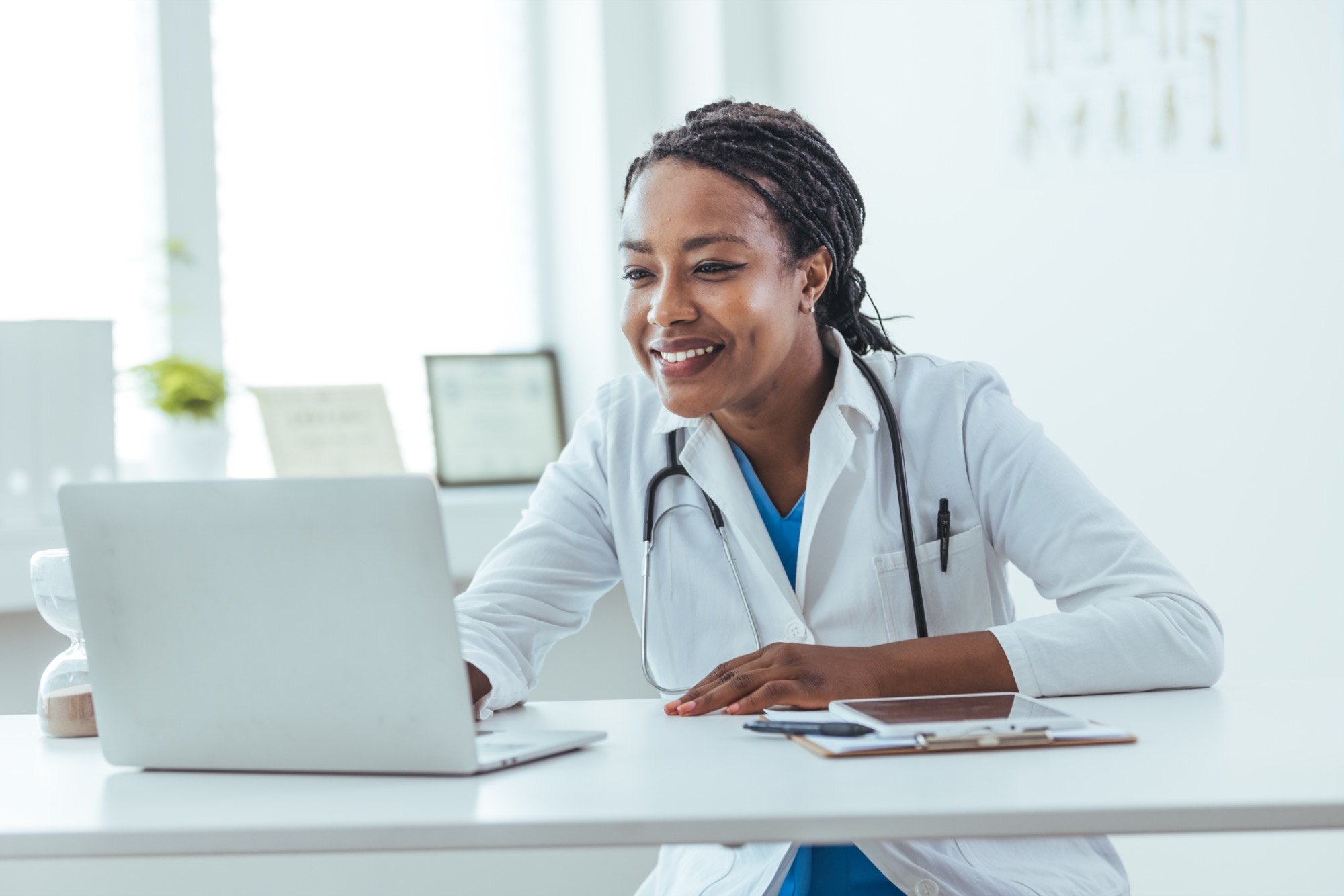 Female doctor with a stethoscope smiling while working on a laptop at her desk