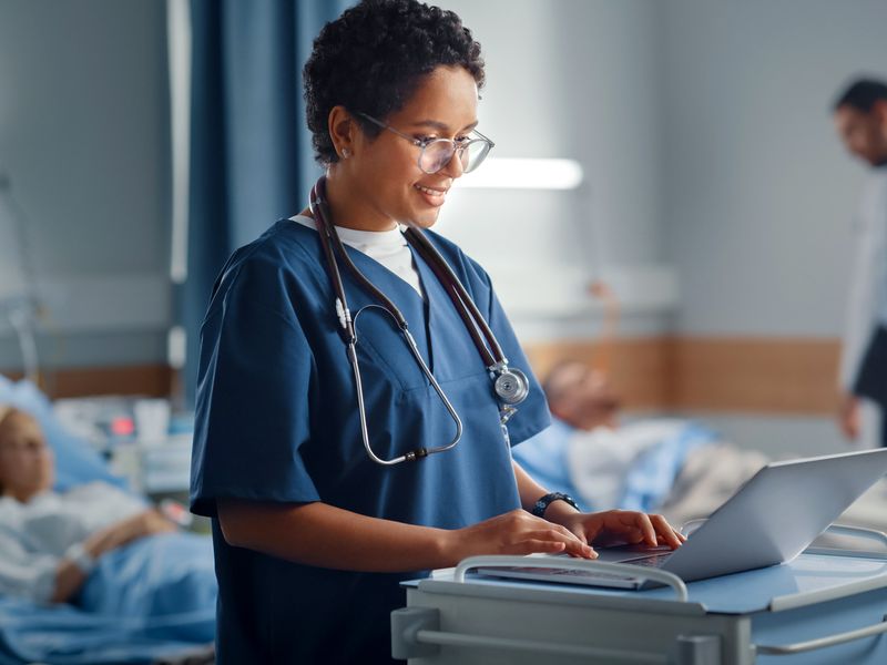 Nurse in scrubs smiling while using a laptop on a medical cart in a hospital ward with patients in the background.