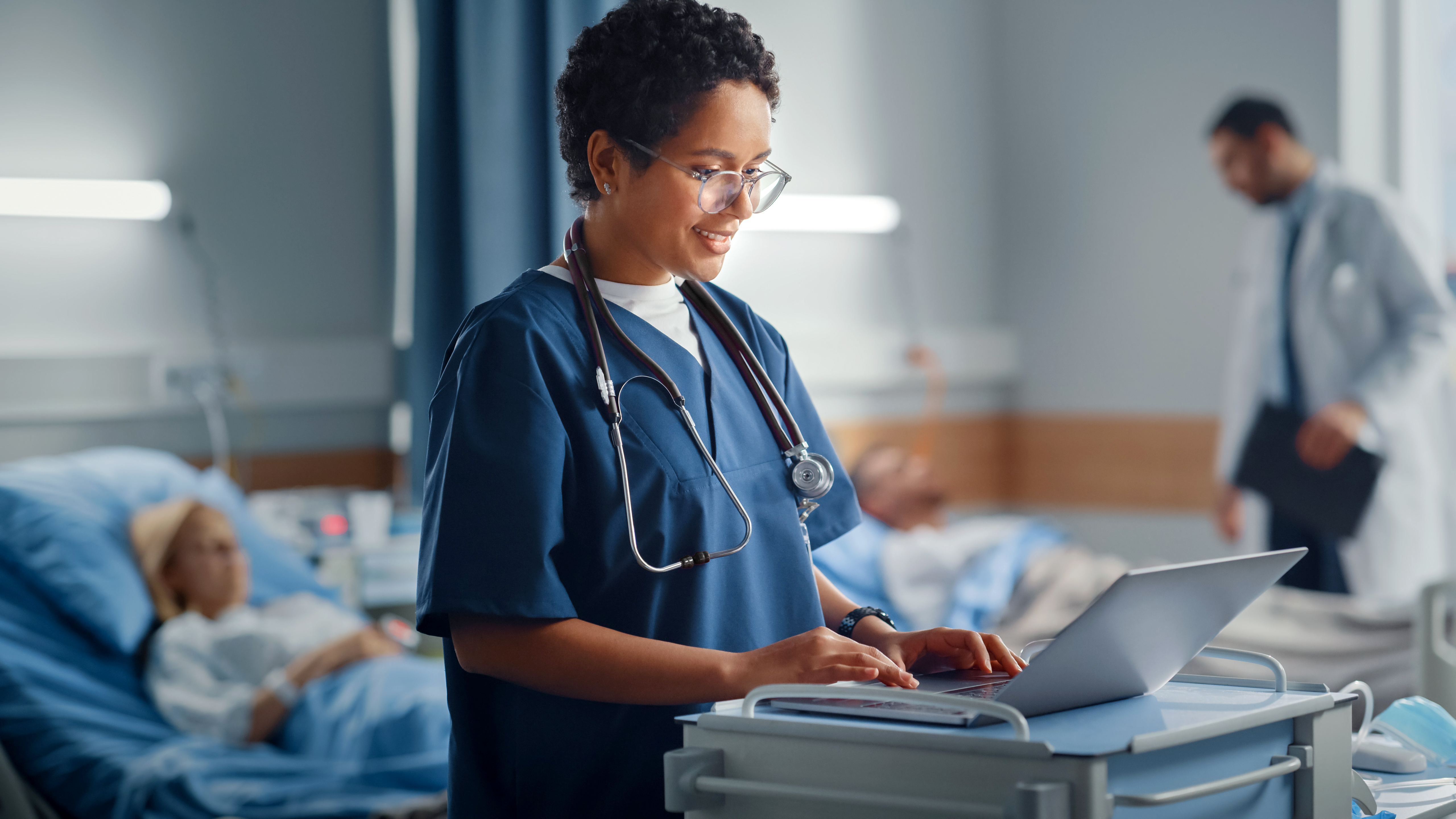 Nurse in scrubs smiling while using a laptop on a medical cart in a hospital ward with patients in the background.