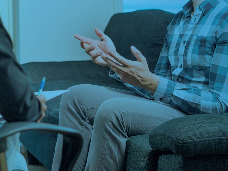 Person sitting on a couch gesturing during a counseling or therapy session, with a blue color overlay.