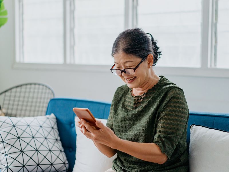 Smiling senior woman wearing glasses using a smartphone while sitting on a couch at home.