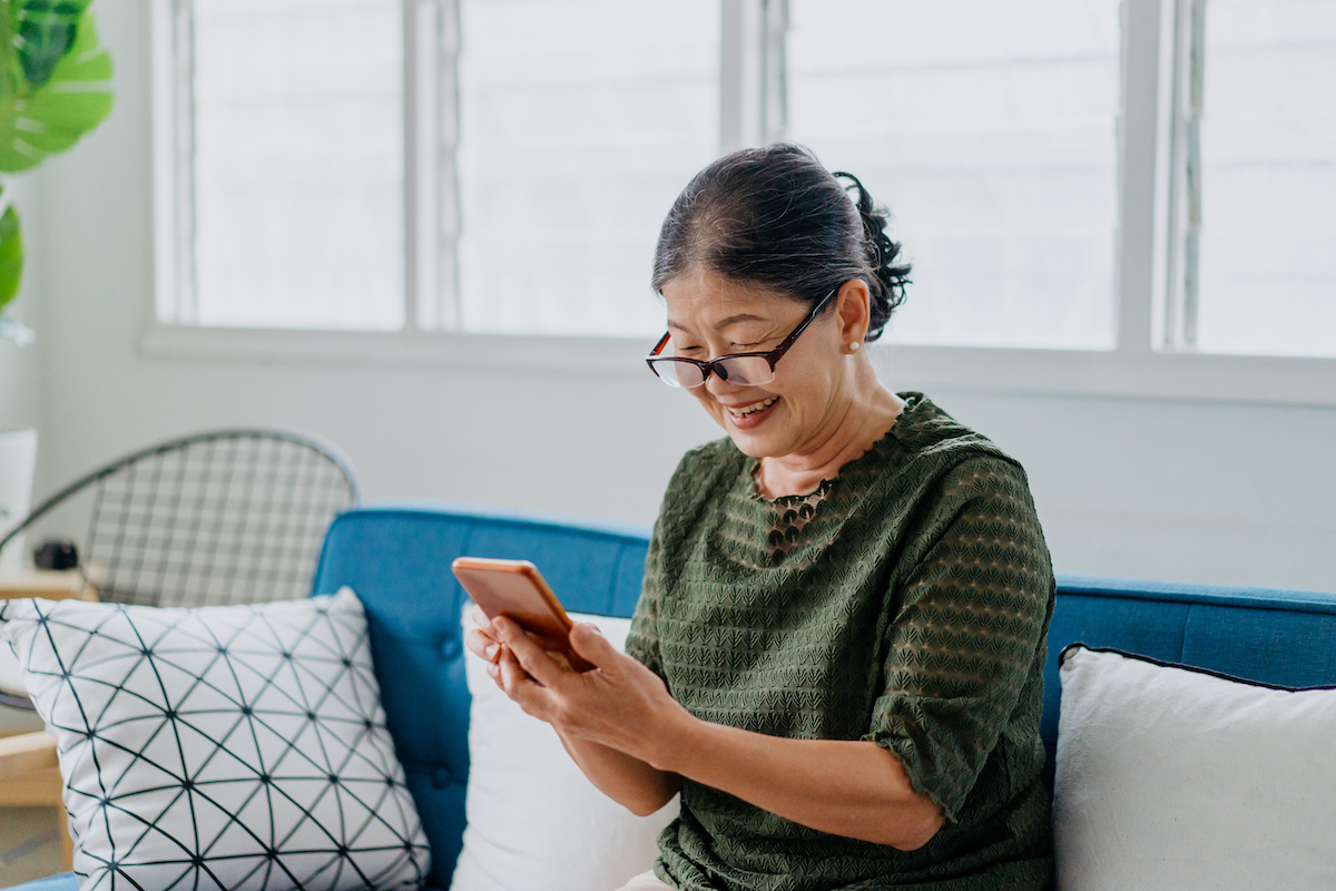 Smiling senior woman wearing glasses using a smartphone while sitting on a couch at home.