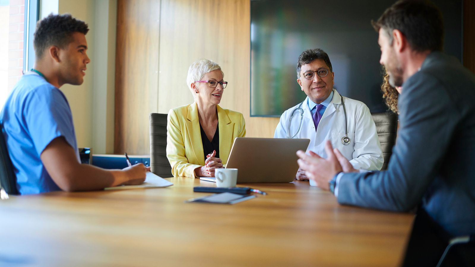 Healthcare professionals and administrators in a conference room meeting, including a doctor in a lab coat and a nurse in scrubs.