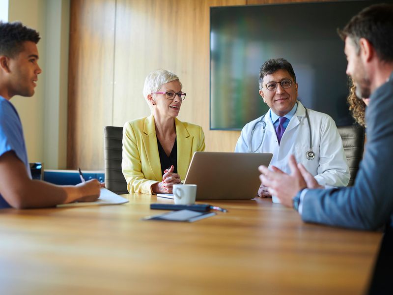 Healthcare professionals and administrators in a conference room meeting, including a doctor in a lab coat and a nurse in scrubs.