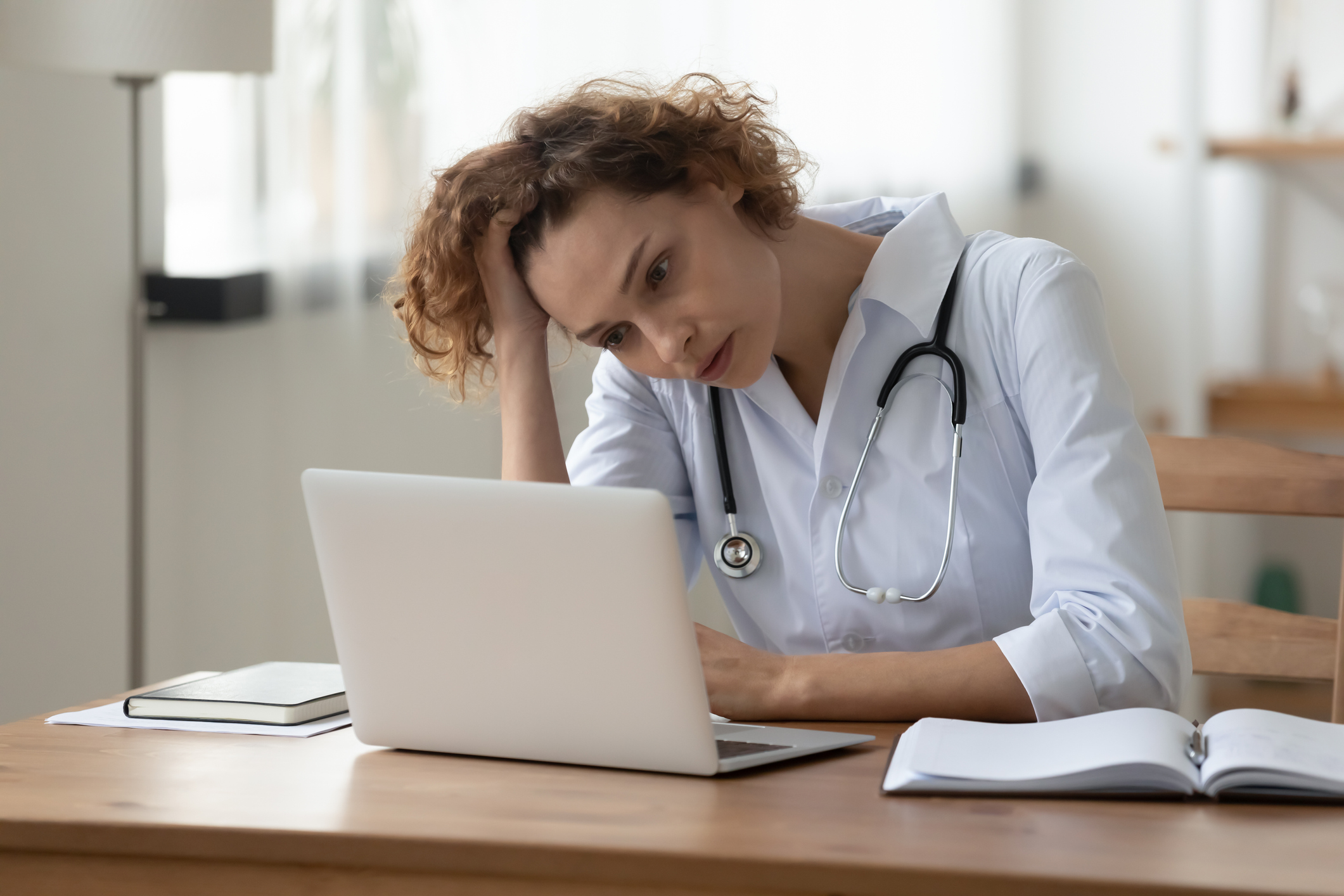 Female doctor with stethoscope looking stressed while reviewing information on a laptop at her desk.