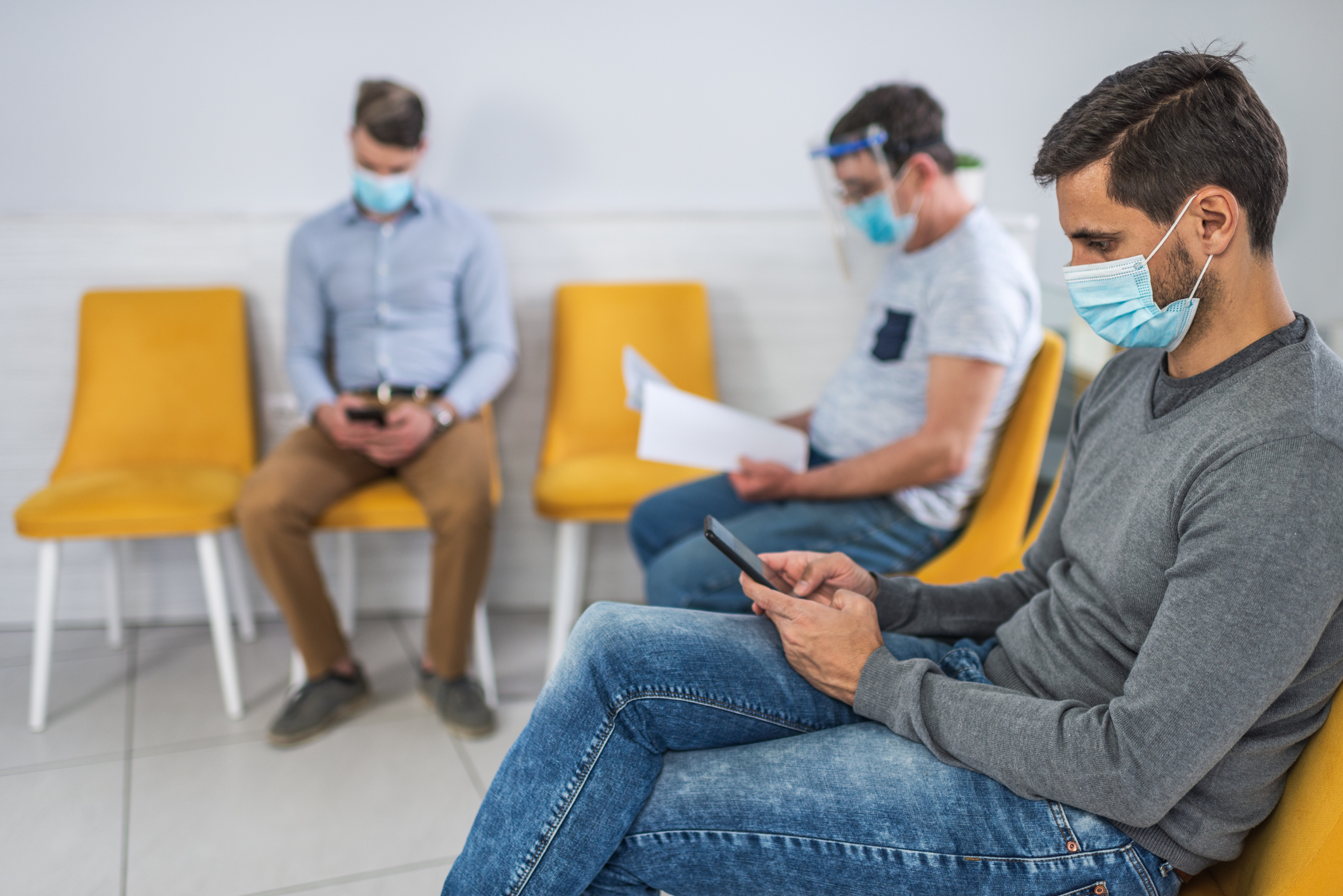 Patients wearing face masks sitting socially distanced in a medical waiting room, using smartphones.