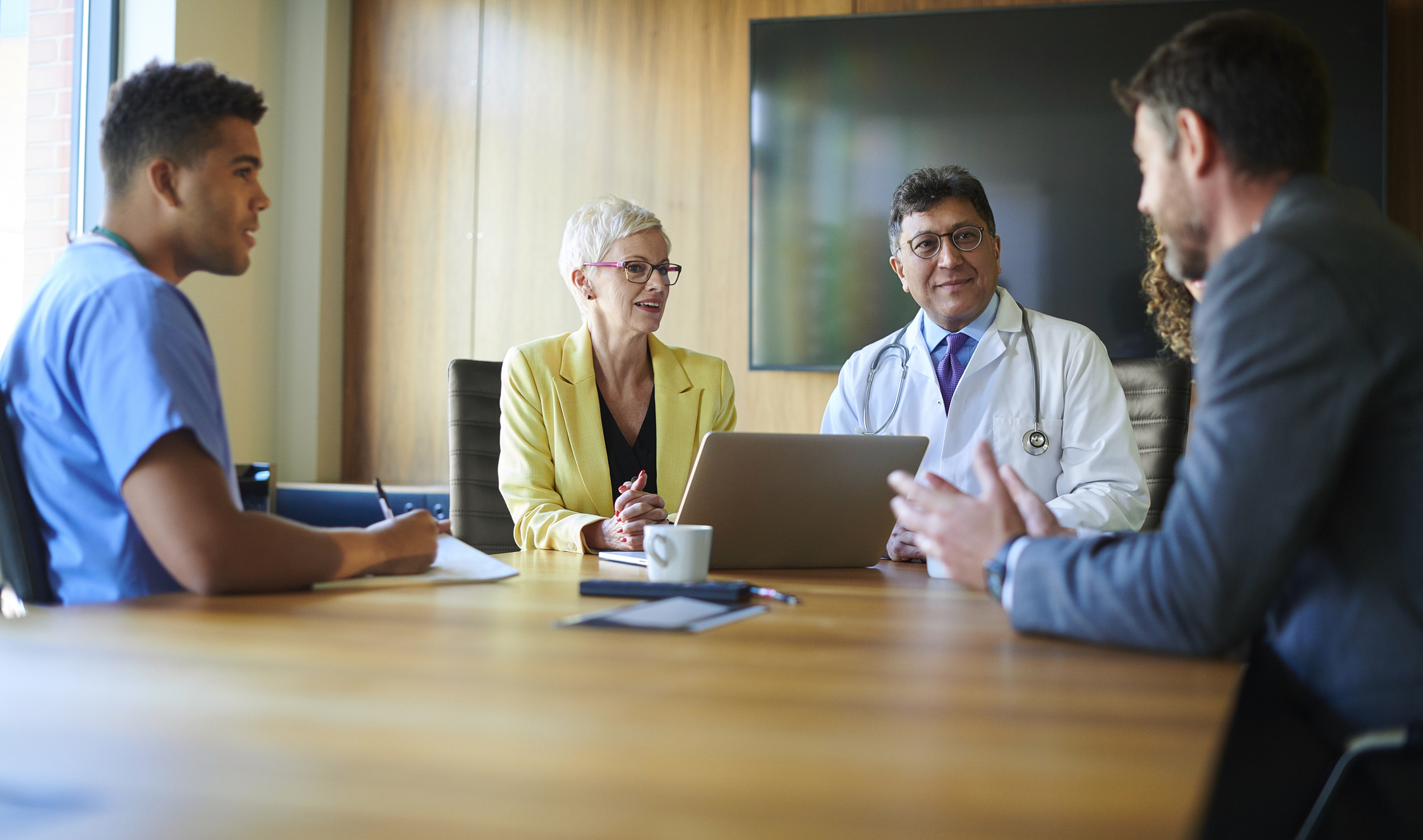 Healthcare professionals and administrators in a conference room meeting, including a doctor in a lab coat and a nurse in scrubs.