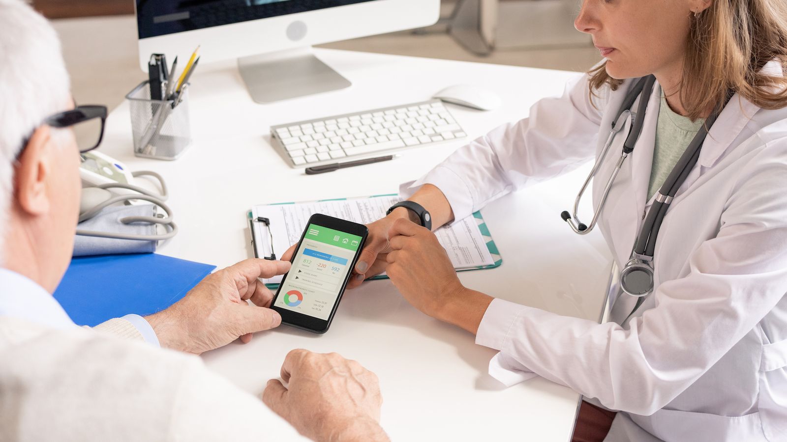 Doctor and elderly patient reviewing health data on a smartphone together at a desk with a computer monitor.
