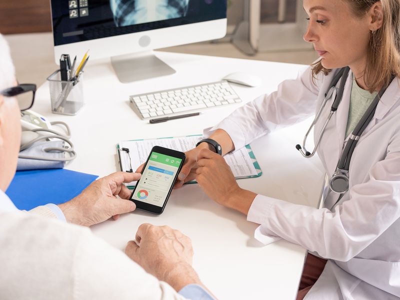 Doctor and elderly patient reviewing health data on a smartphone together at a desk with a computer monitor.
