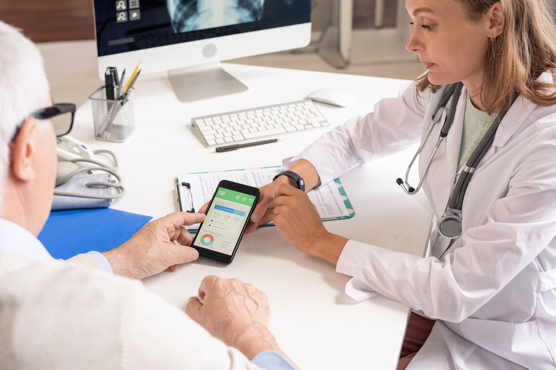 Doctor and elderly patient reviewing health data on a smartphone together at a desk with a computer monitor.