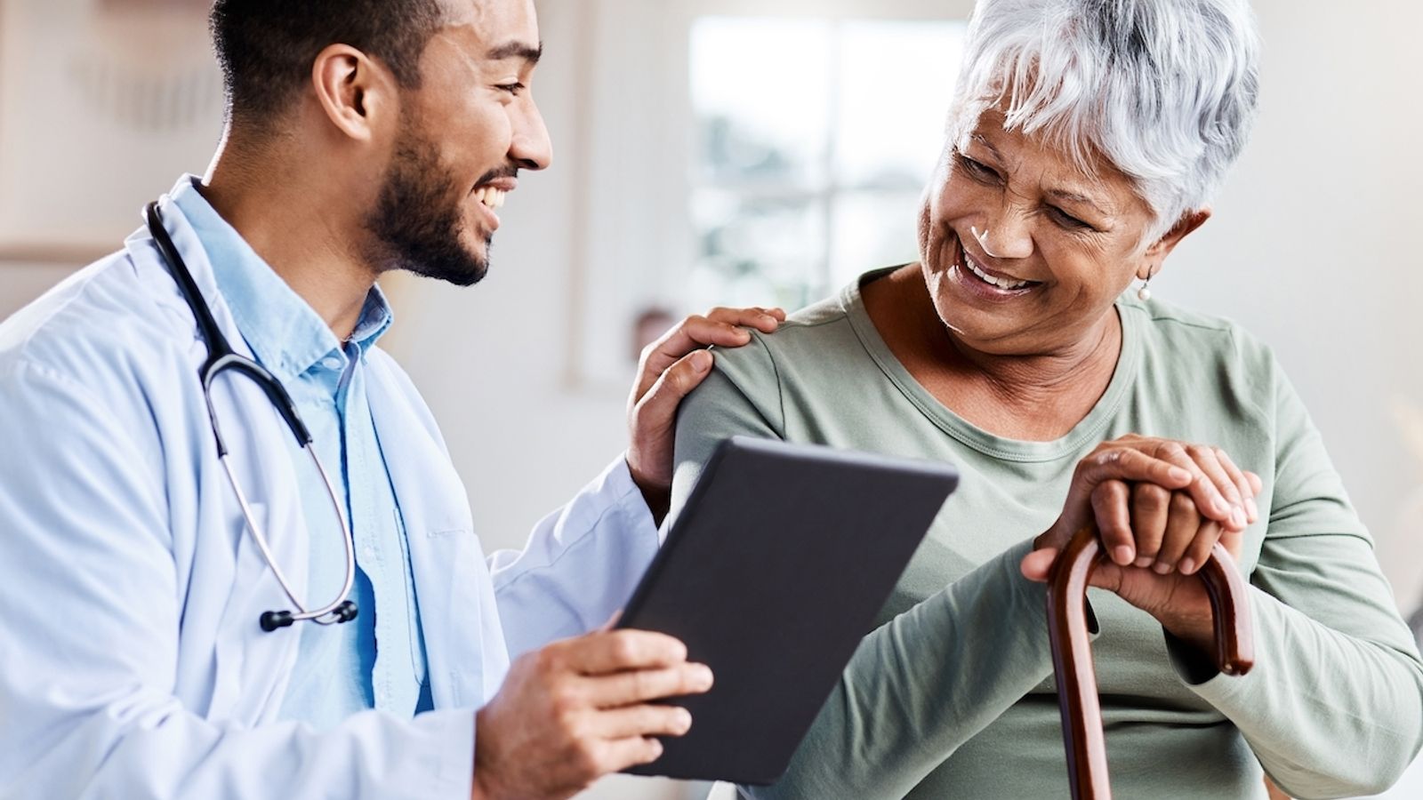 Doctor smiling and showing a tablet to a senior patient with a walking cane during a home visit.