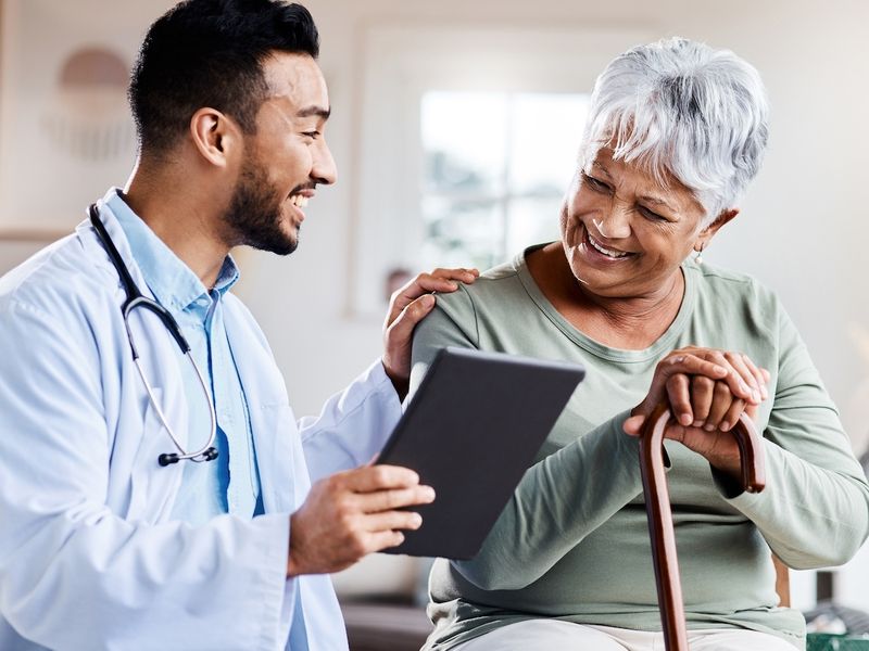 Doctor smiling and showing a tablet to a senior patient with a walking cane during a home visit.