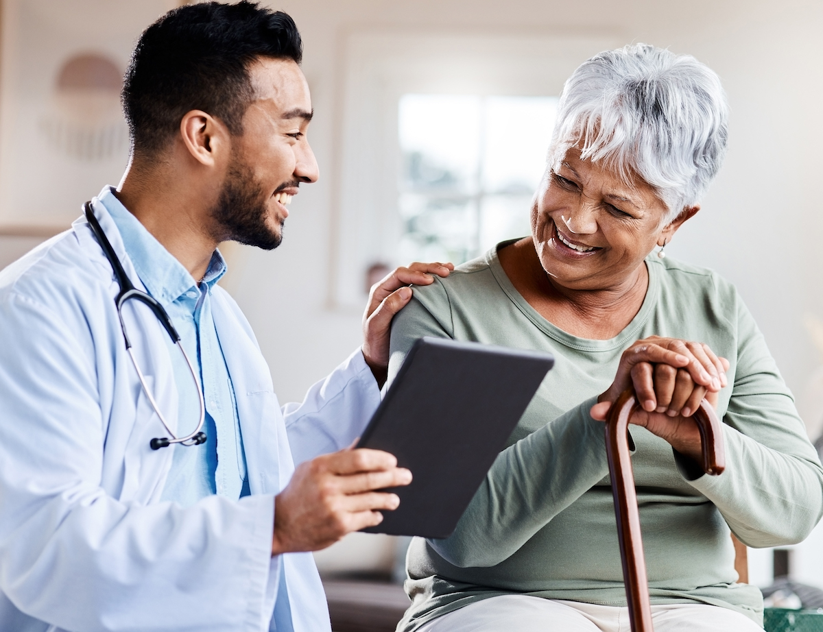 Doctor smiling and showing a tablet to a senior patient with a walking cane during a home visit.