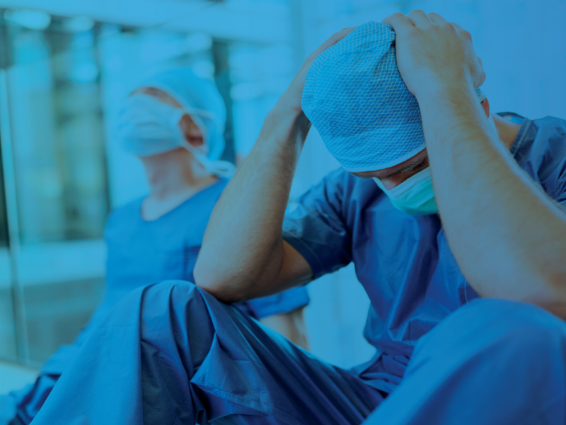 Healthcare worker in scrubs sitting with head in hands, conveying stress or burnout, with a dark blue overlay.