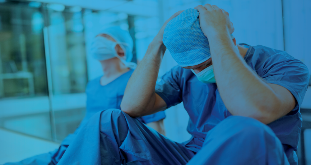 Healthcare worker in scrubs sitting with head in hands, conveying stress or burnout, with a dark blue overlay.