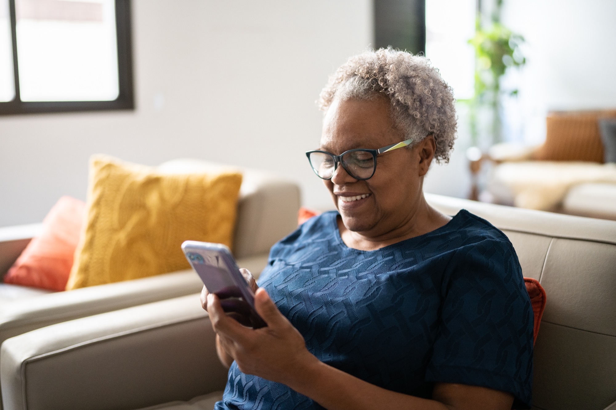 Senior woman smiling while using a smartphone on her living room couch