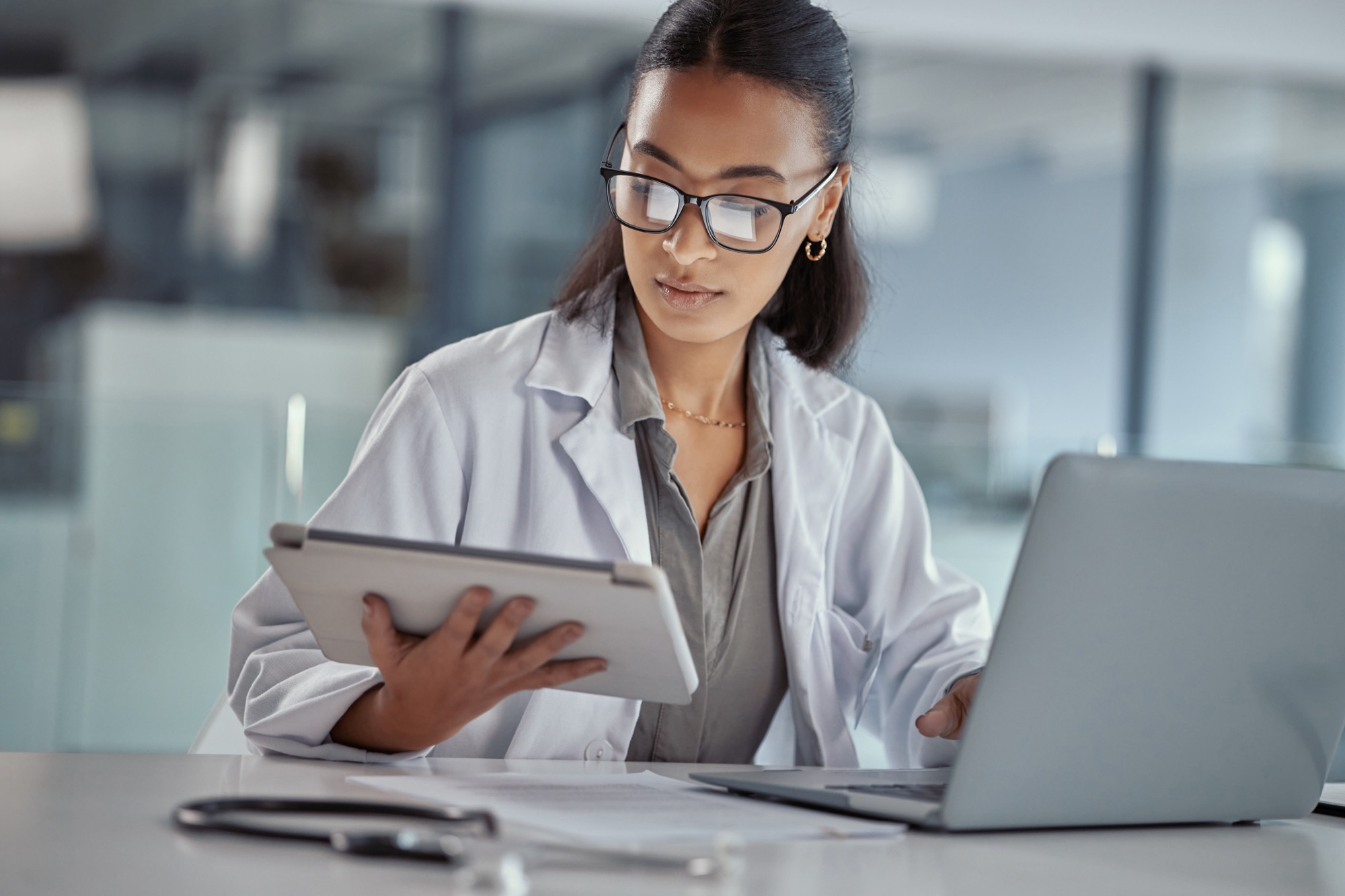 Female doctor in a lab coat reviewing patient data on a digital tablet alongside a laptop